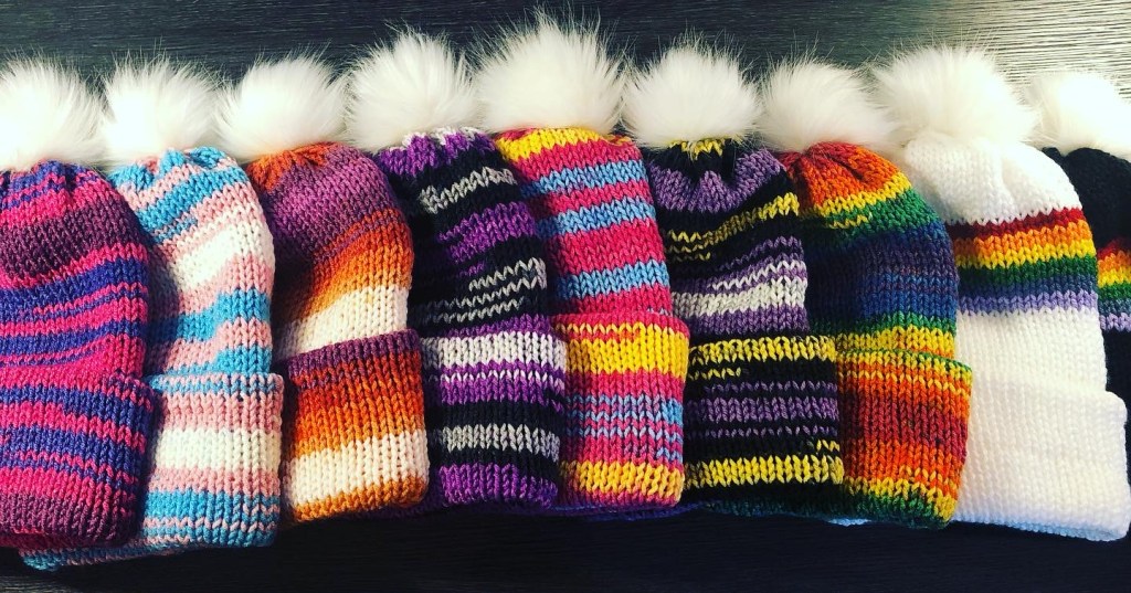 Nine hand-knitted, 2SLGBTQ+ color themed hats laid out on a black table top.