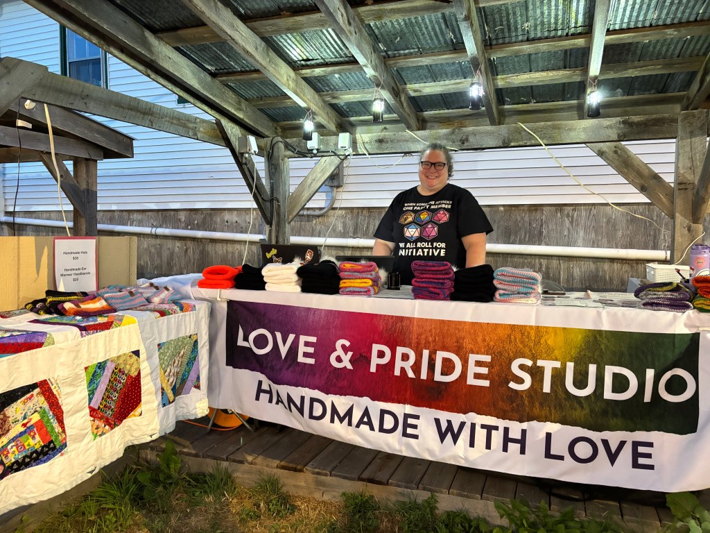 Brigit smiling and standing behind market table with Love & Pride Studio banner. Knitted products and custom magnet maker are on the table.